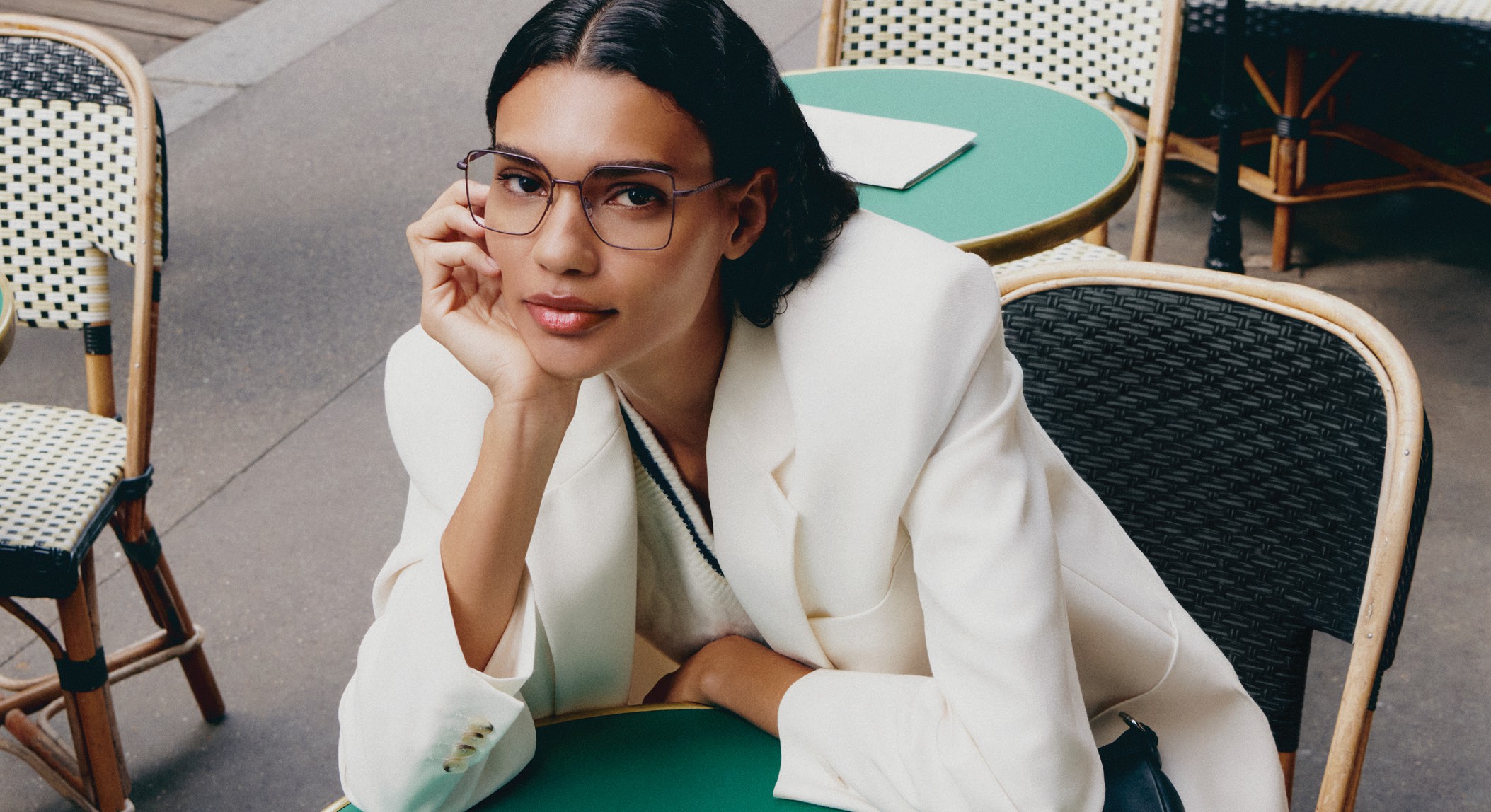Woman wearing Lacoste L2316 optical glasses in a white blazer seated at a mint green café table in a modern Parisian setting