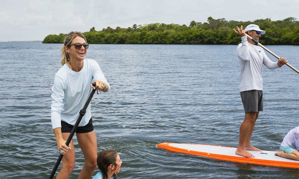Sarah Scott portant des lunettes de soleil sur une planche à pagaie sur une eau calme avec KC Scott sur une planche à pagaie en arrière-plan près d'une côte de mangroves