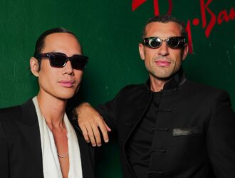 Two people wearing distinctive Ray-Ban sunglasses pose against a green wall with Ray-Ban branding visible in red lettering