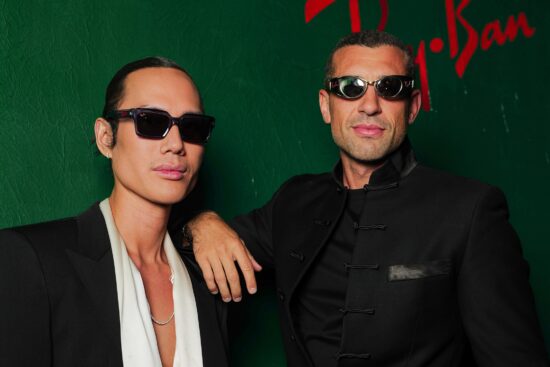 Deux personnes portant des lunettes de soleil Ray-Ban distinctives posent devant un mur vert avec le logo Ray-Ban visible en lettres rouges
