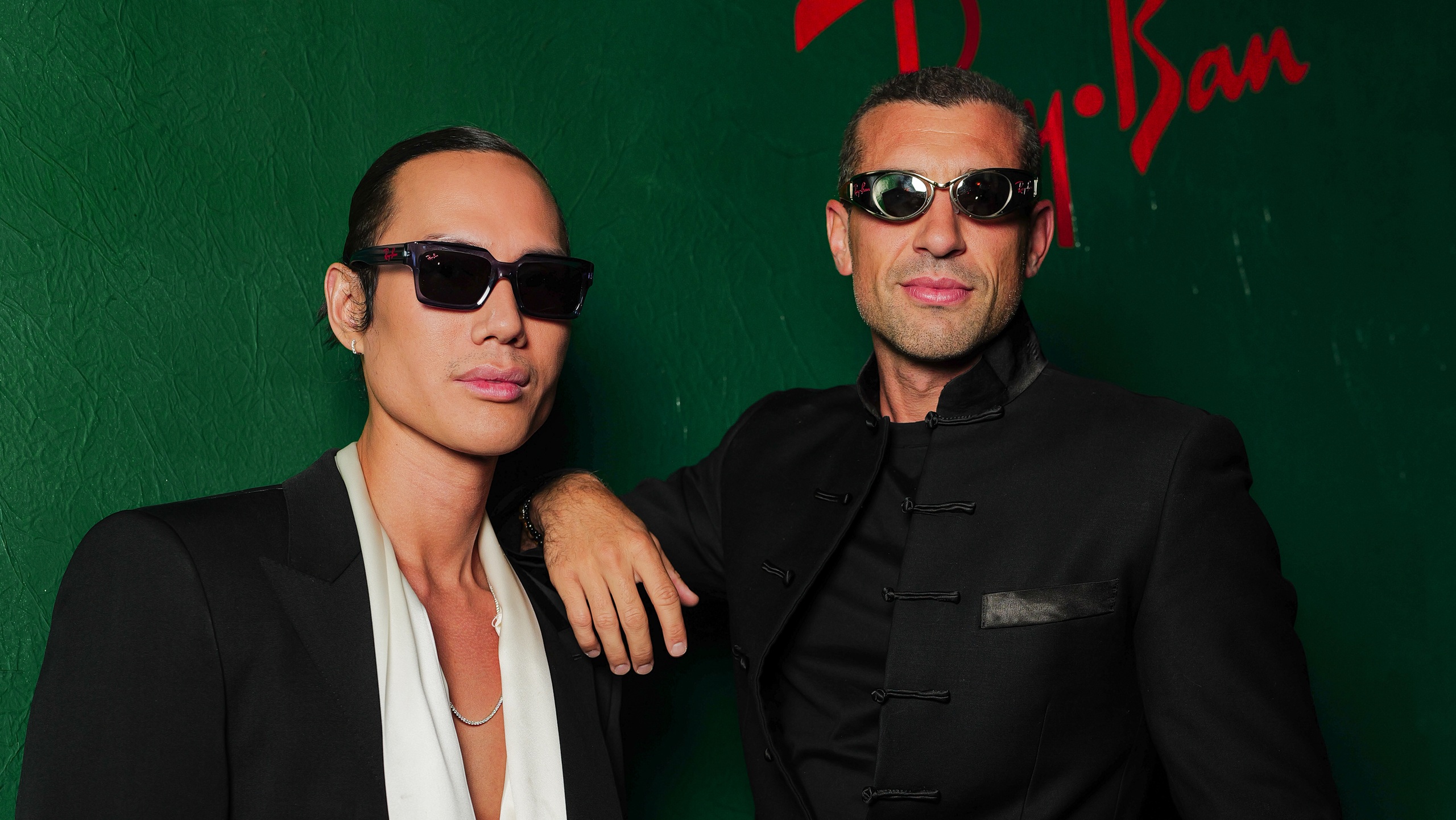 Deux personnes portant des lunettes de soleil Ray-Ban distinctives posent devant un mur vert avec le logo Ray-Ban visible en lettres rouges