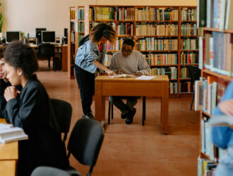 Diverse group of students studying in a bright library with bookshelves, some working on laptops at desks while others read books together at tables