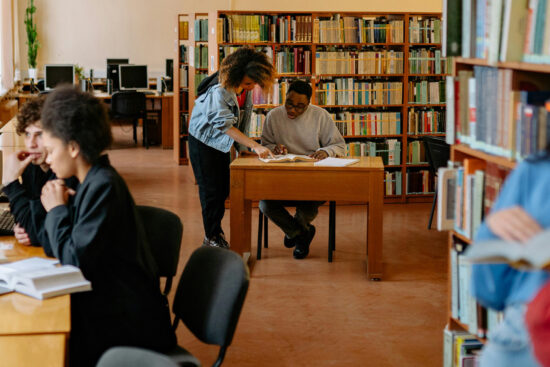 Diverse group of students studying in a bright library with bookshelves, some working on laptops at desks while others read books together at tables