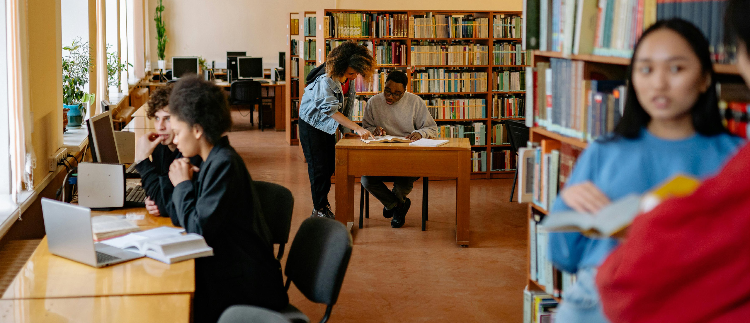 Diverse group of students studying in a bright library with bookshelves, some working on laptops at desks while others read books together at tables