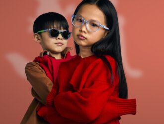Two children wearing Ray-Ban Disney Zootopia 2 collection glasses against a coral pink gradient background; one child wears transparent blue sunglasses with green accents, the other wears transparent blue optical frames with orange details