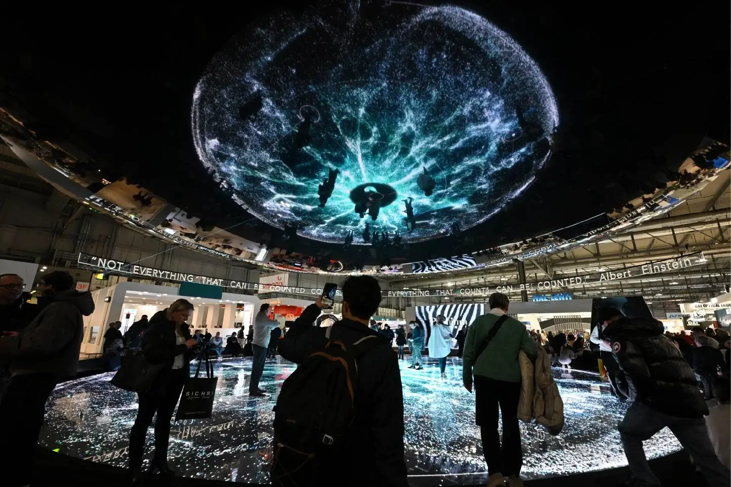 Based on the image, here's an appropriate alt text: "Large circular LED screen display showing water splash effects in turquoise and white above crowds at MIDO eyewear trade show, with Einstein quote visible on surrounding walls