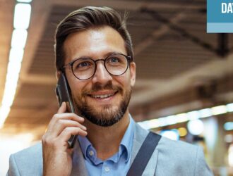 Un homme souriant portant des lunettes et un complet parle sur un téléphone mobile tout en tenant une tasse de café en carton, debout dans un espace intérieur très éclairé qui semble être une station de transport en commun. L'image comporte une boîte bleue et blanche dans le coin supérieur droit avec le texte « DAYNAMIC PRO ».