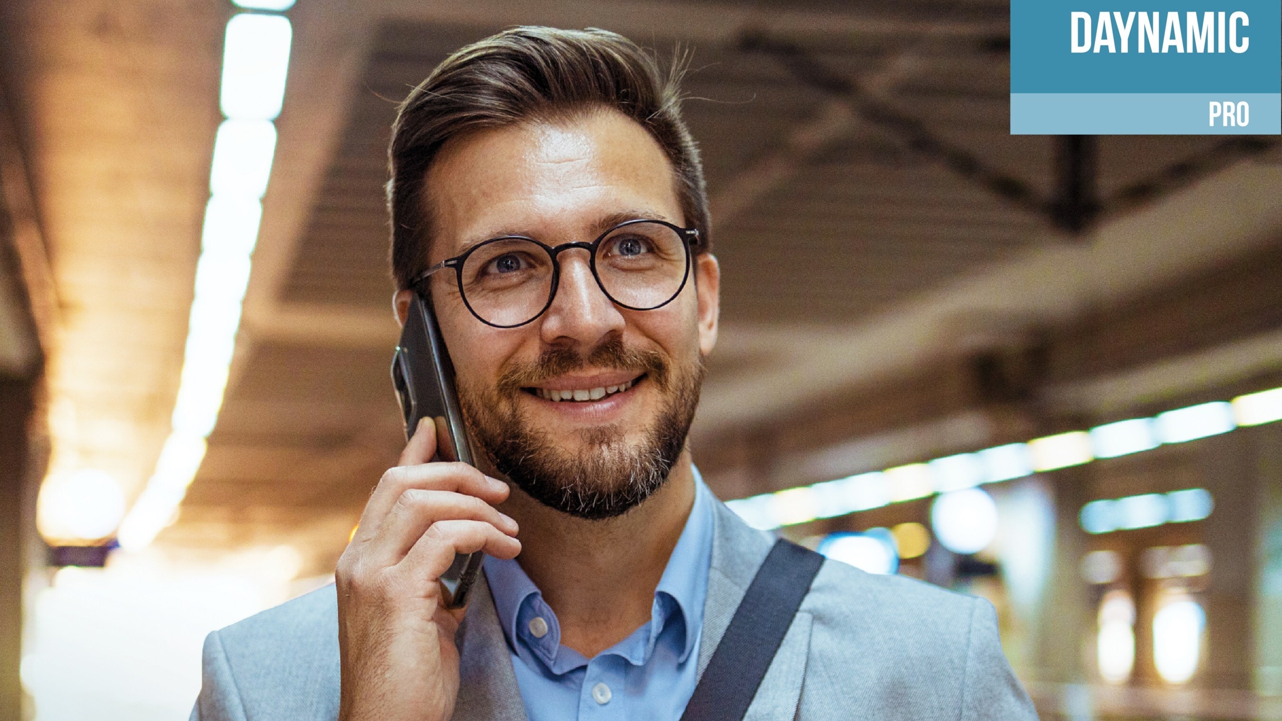Un homme souriant portant des lunettes et un complet parle sur un téléphone mobile tout en tenant une tasse de café en carton, debout dans un espace intérieur très éclairé qui semble être une station de transport en commun. L'image comporte une boîte bleue et blanche dans le coin supérieur droit avec le texte « DAYNAMIC PRO ».