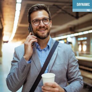 Un homme souriant portant des lunettes et un complet parle sur un téléphone mobile tout en tenant une tasse de café en carton, debout dans un espace intérieur très éclairé qui semble être une station de transport en commun. L'image comporte une boîte bleue et blanche dans le coin supérieur droit avec le texte « DAYNAMIC PRO ».
