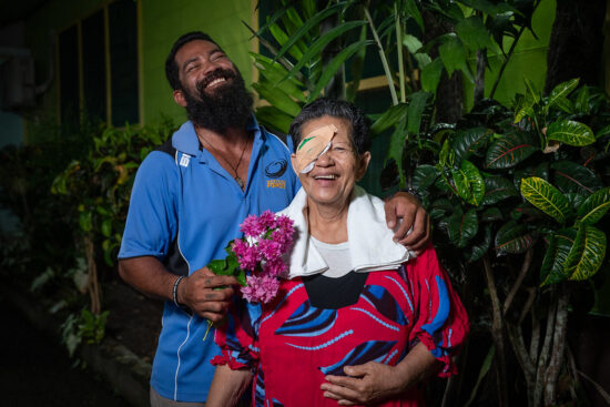 Portrait joyeux post-opératoire montrant une femme âgée avec un pansement oculaire et son fils adulte s'enlaçant dans un jardin tropical, tous deux souriant chaleureusement alors qu'il tient des fleurs roses vives