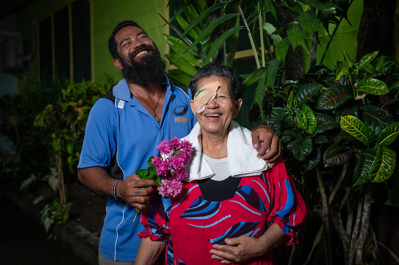 A joyful post-surgery portrait showing an older woman with an eye patch and her adult son embracing in a tropical garden setting, both smiling warmly as he holds bright pink flowers