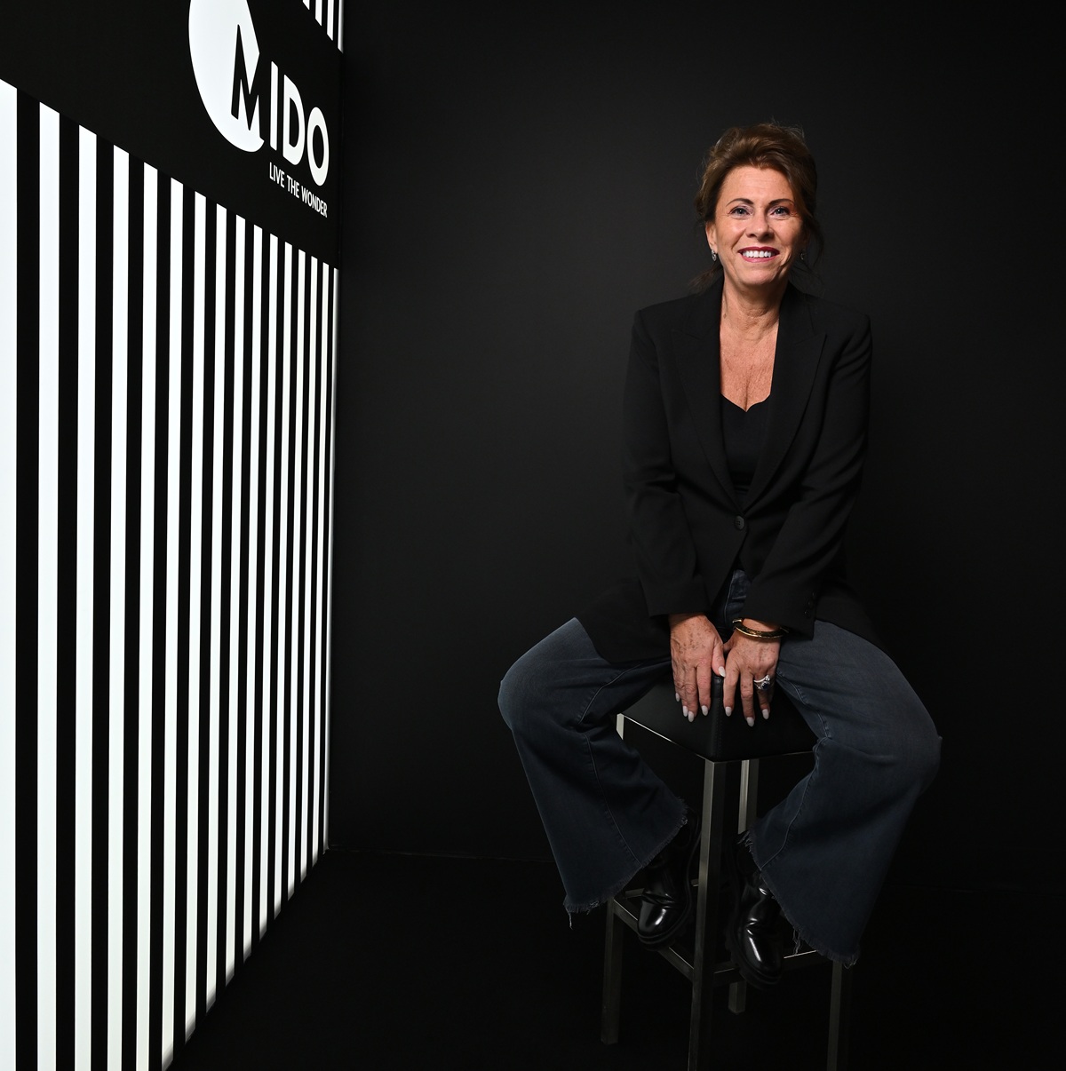 Lorraine Berton, President of ANFAO and MIDO, seated on a stool in elegant black business attire against a dark backdrop featuring the MIDO logo with vertical white stripes