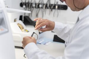 Gros plan d'un professionnel de la vue en blouse blanche travaillant sur les verres d'une paire de lunettes, avec des outils optiques et une machine de taillage visibles en arrière-plan.
