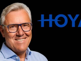 Portrait professionnel d' Oliver Fischbach souriant portant des lunettes à monture bleue et une chemise bleu clair sur fond noir
