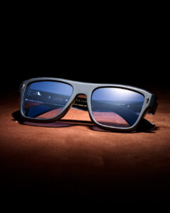 OTH eyewear optical frame in matte grey acetate with blue-tinted lenses and metal detailing on temples, displayed on warm-toned textured surface