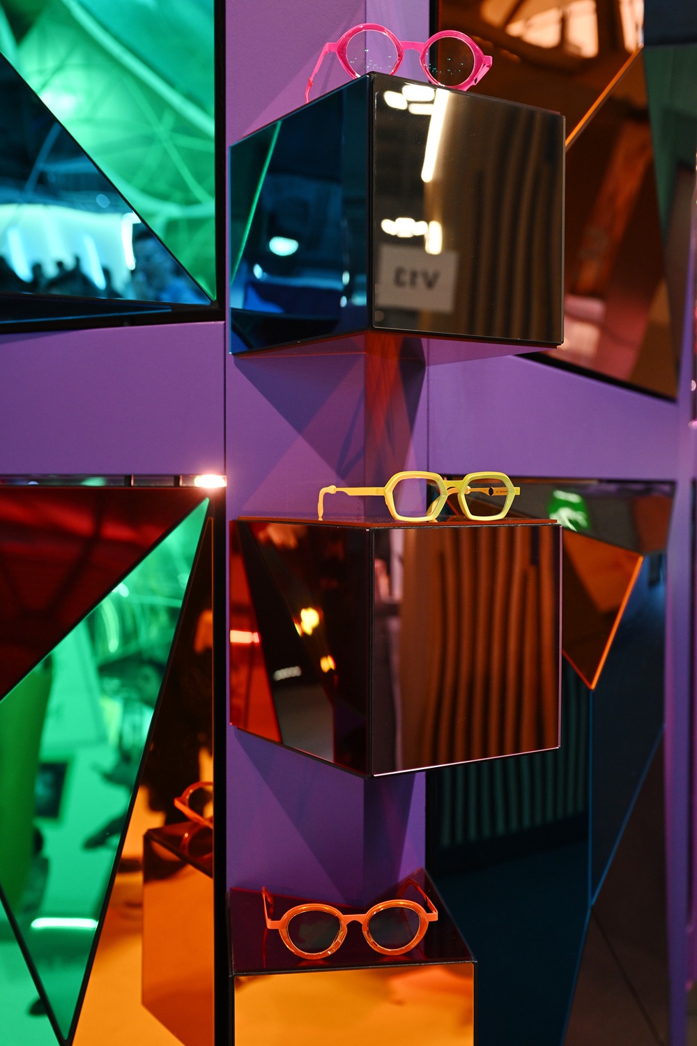 Three pairs of brightly colored, contemporary eyewear (pink, yellow-green, and orange) displayed on a mirrored purple wall structure with multicolored, angular reflective panels at an exhibition.