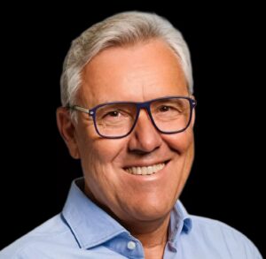 Professional headshot of a smiling executive wearing blue-framed glasses and a light blue shirt against a black background