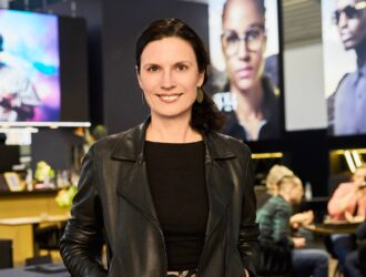 Professional woman in black leather jacket standing in modern optical retail space with eyewear displays in background