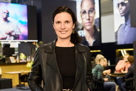 Professional woman in black leather jacket standing in modern optical retail space with eyewear displays in background
