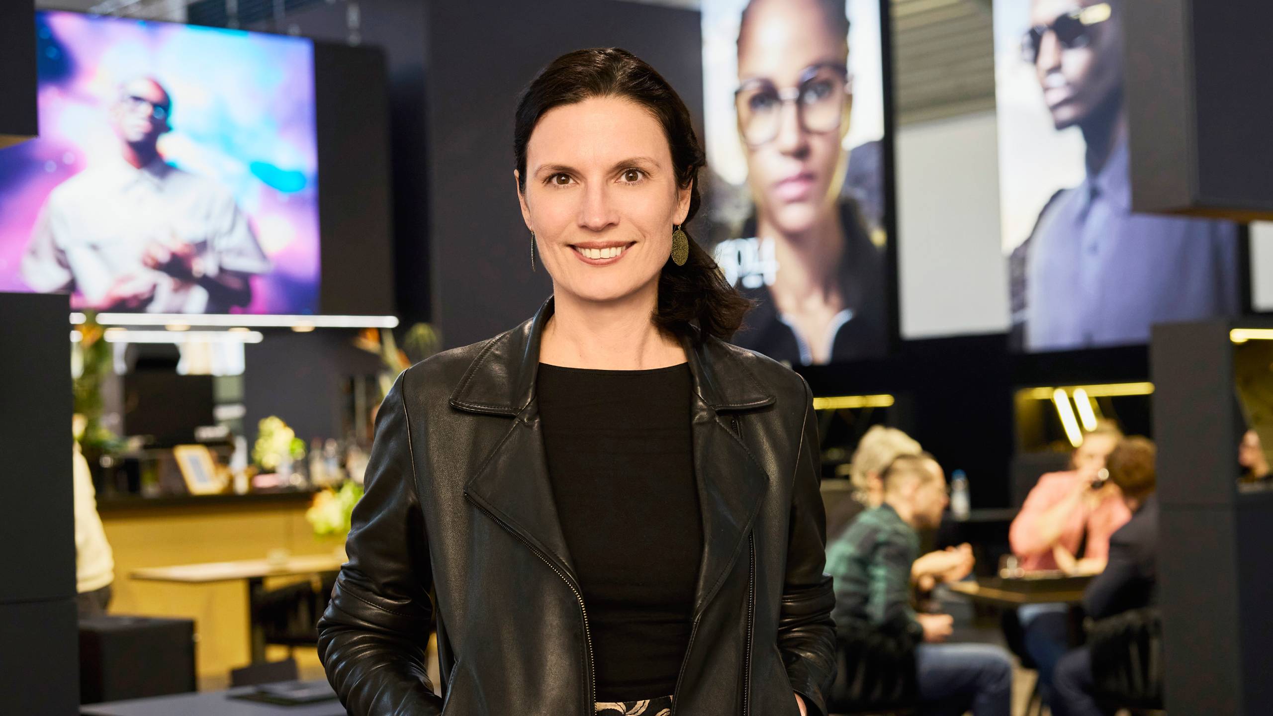 Professional woman in black leather jacket standing in modern optical retail space with eyewear displays in background
