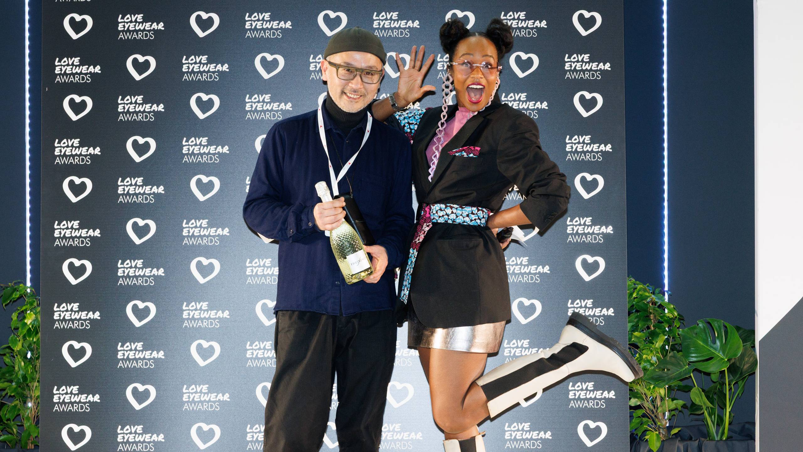 Deux personnes posant de manière enjouée devant un mur de logos « Love Eyewear Awards » ; l'une tient une bouteille de champagne tandis que l'autre lève une jambe en souriant.