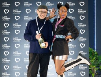 Two people posing playfully in front of a "Love Eyewear Awards" backdrop; one holds a bottle of champagne while the other poses with a raised leg.