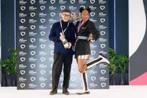 Two people posing playfully in front of a "Love Eyewear Awards" backdrop; one holds a bottle of champagne while the other poses with a raised leg.