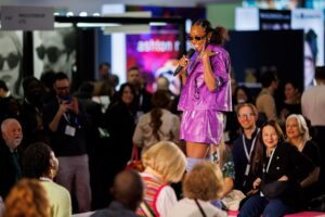 A presenter in a bright purple outfit speaking into a microphone on a stage during the 100% Optical event, surrounded by an audience and exhibition booths.