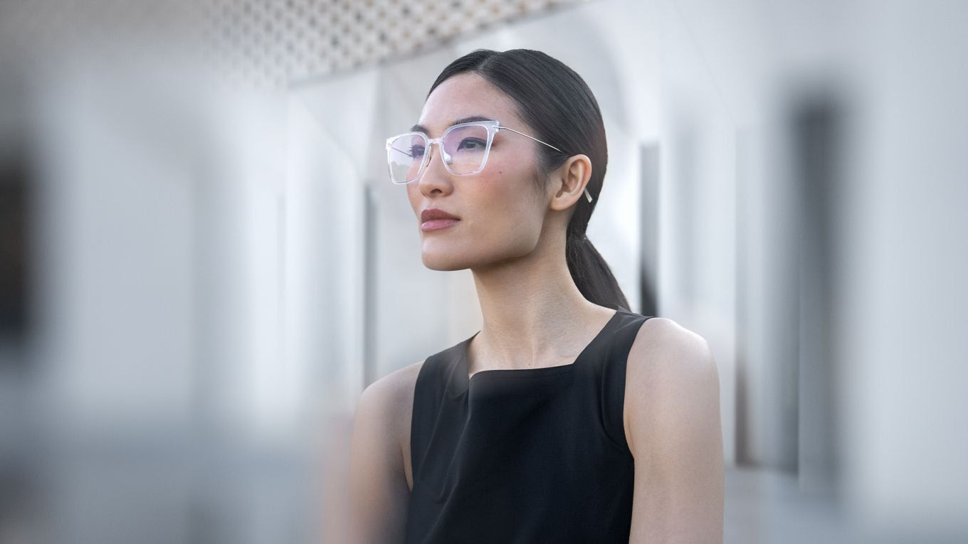 A woman wearing oversized transparent Blackfin Vitra frames with thin metallic temples against a bright, modern background.