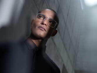 A man wearing clear, square-framed Blackfin Vitra glasses looking upward in a minimalist, architectural setting.