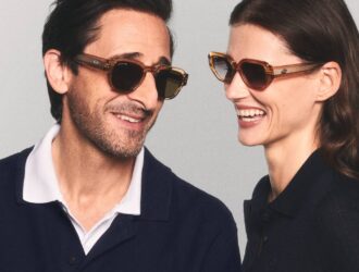 Academy Award-winning actor and Lacoste Global Eyewear Ambassador Adrien Brody smiles alongside a female model in a promotional campaign shot. Both are wearing modern, geometric Lacoste sunglasses with transparent honey-colored frames and dark lenses. They are dressed in classic Lacoste polo shirts, reflecting the brand’s "Unconventional Chic" and heritage style.