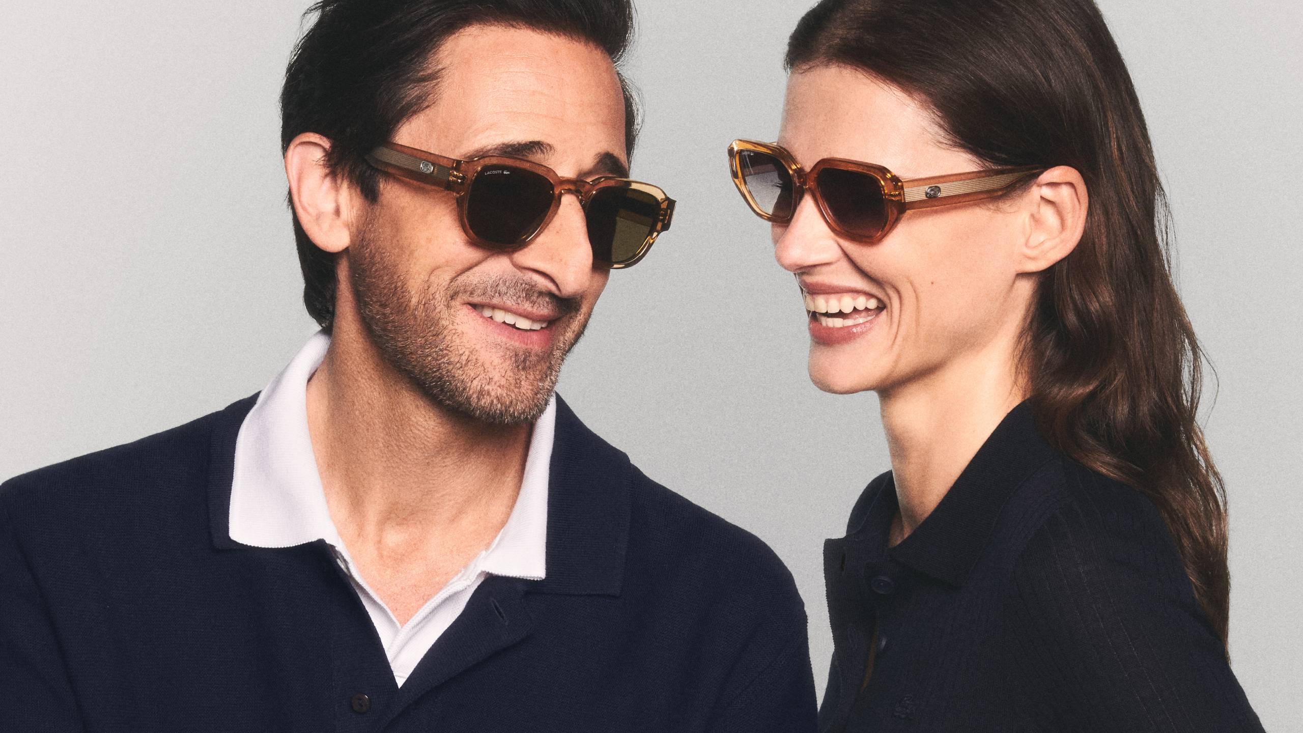 Academy Award-winning actor and Lacoste Global Eyewear Ambassador Adrien Brody smiles alongside a female model in a promotional campaign shot. Both are wearing modern, geometric Lacoste sunglasses with transparent honey-colored frames and dark lenses. They are dressed in classic Lacoste polo shirts, reflecting the brand’s "Unconventional Chic" and heritage style.