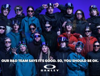 Group photo of Oakley's R&D team wearing the AURA Collection eyewear, goggles, and helmets with iridescent colourshift finish against a purple background, with text reading 'Our R&D team says it's good. So, you should be ok.