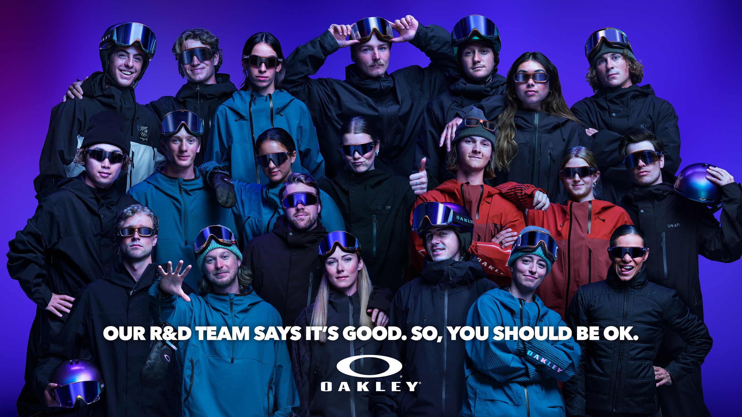 Group photo of Oakley's R&D team wearing the AURA Collection eyewear, goggles, and helmets with iridescent colourshift finish against a purple background, with text reading 'Our R&D team says it's good. So, you should be ok.