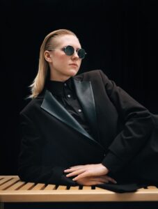 Model in round metallic sunglasses from Ørgreen's Imaginary Lines collection, wearing black oversized coat, leaning on wooden surface against black backdrop