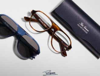 A flat lay of two pairs of Oliver Peoples Paul Newman aviator-style glasses—one blue sunglass and one tortoise optical frame—resting next to a navy blue protective case