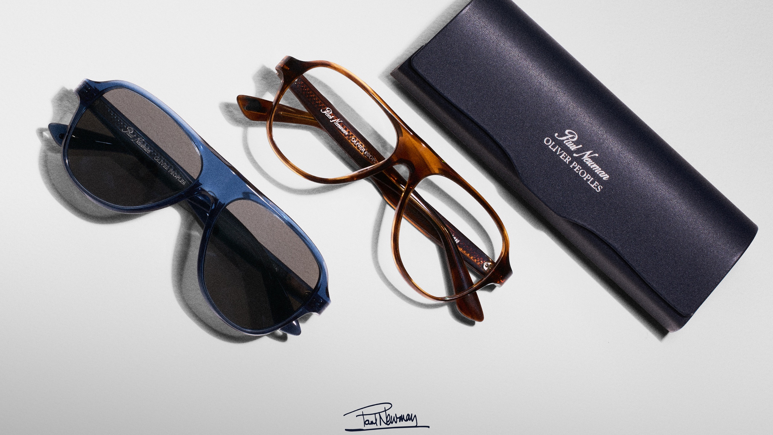 A flat lay of two pairs of Oliver Peoples Paul Newman aviator-style glasses—one blue sunglass and one tortoise optical frame—resting next to a navy blue protective case