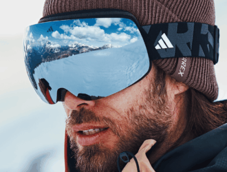 Skier wearing Adidas snow goggles with blue mirrored lens and black strap, brown knit beanie, mountain landscape reflected in lens