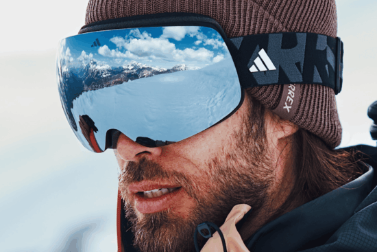 Skier wearing Adidas snow goggles with blue mirrored lens and black strap, brown knit beanie, mountain landscape reflected in lens
