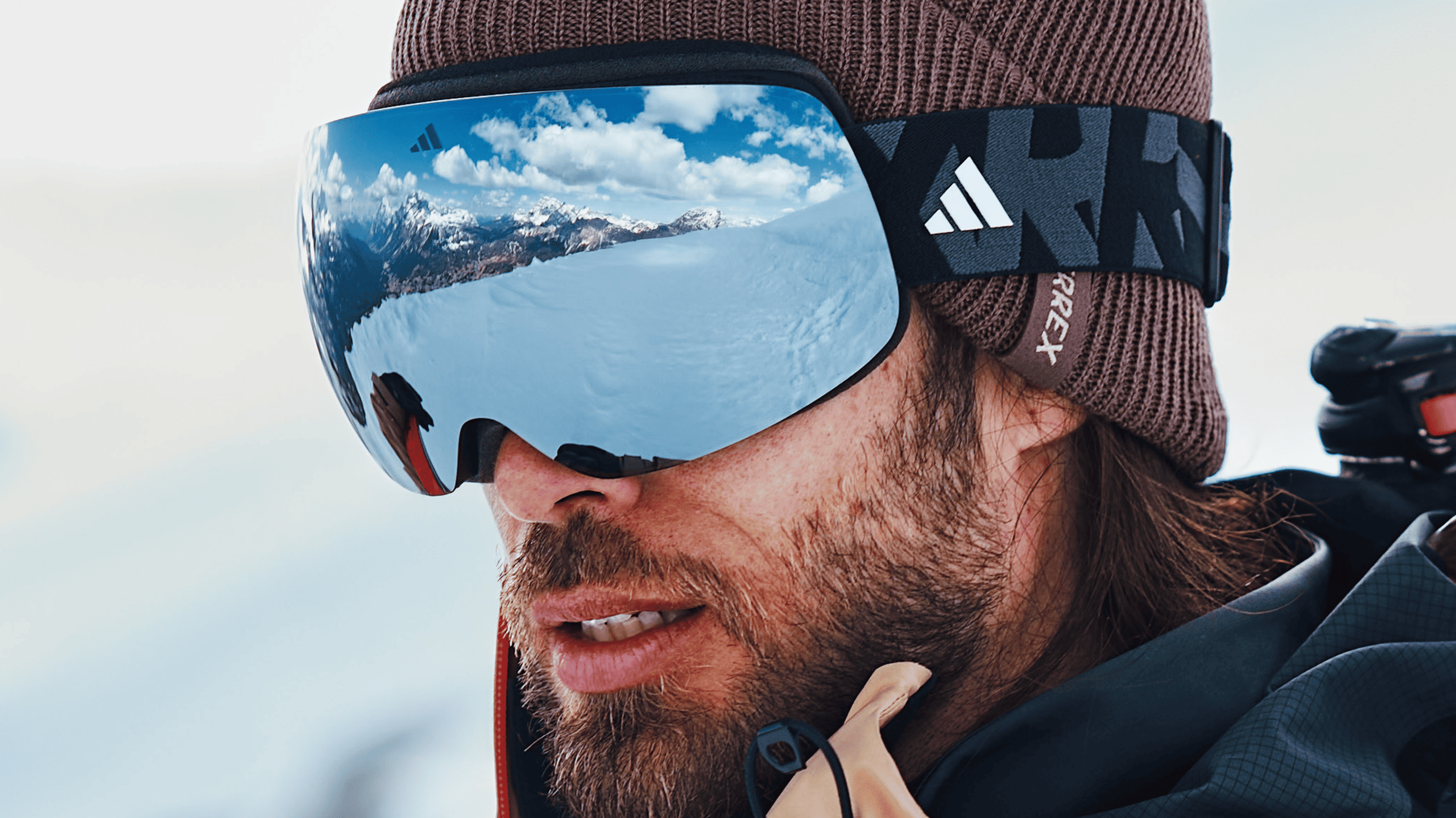Skier wearing Adidas snow goggles with blue mirrored lens and black strap, brown knit beanie, mountain landscape reflected in lens