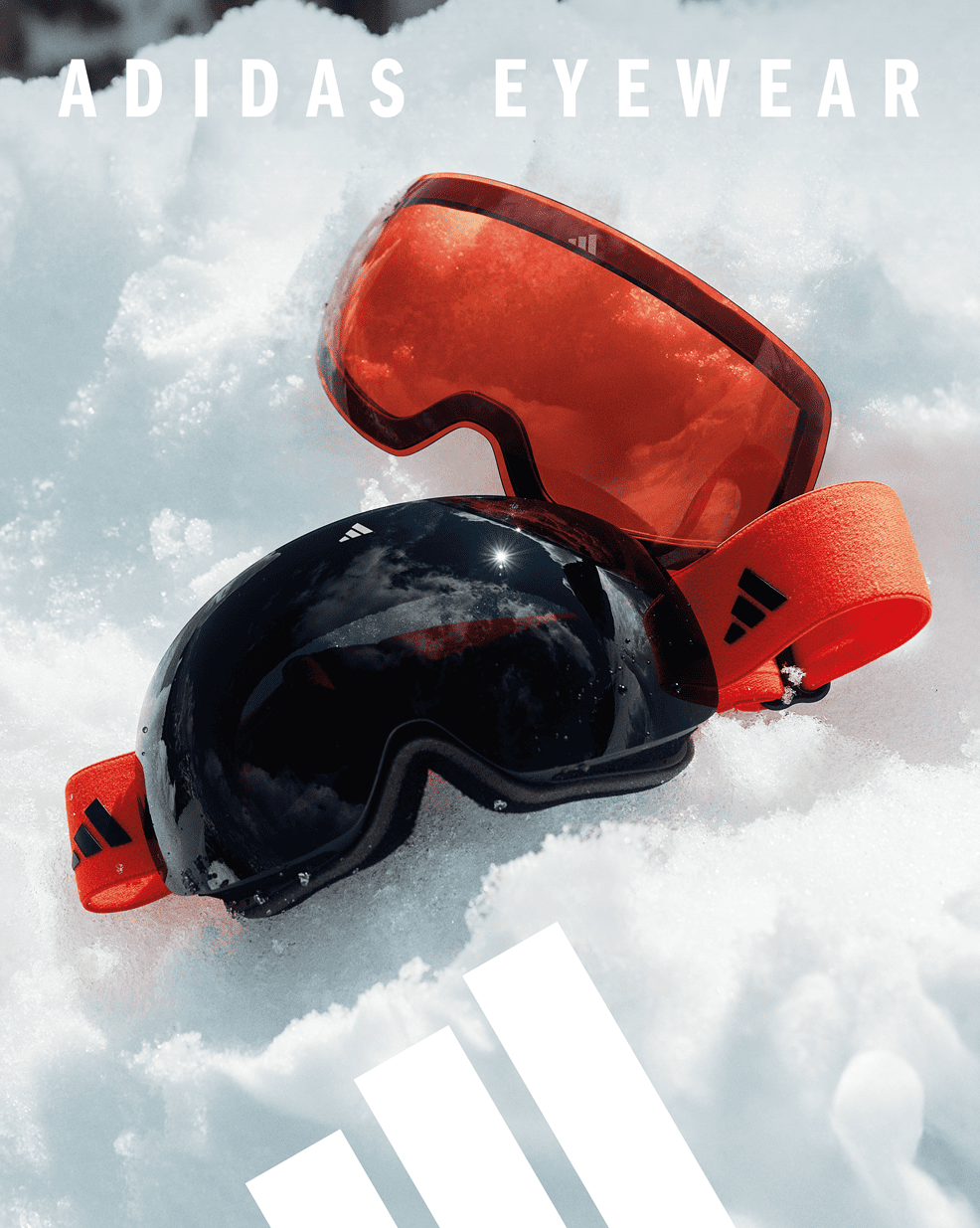 Two pairs of Adidas snow goggles in snow - one black with dark lens and one orange/red with amber lens, both featuring bright orange straps with Adidas logo