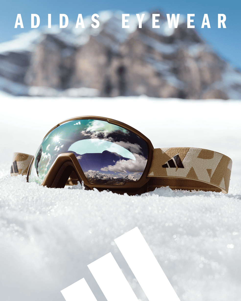 Adidas snow goggles with brown frame and camouflage strap resting in snow with reflective blue-tinted lens showing mountain scenery