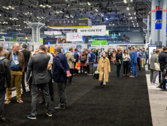 A busy eyewear trade show floor with crowds of people walking between branded exhibit booths like Capri Optics and New York Eye.