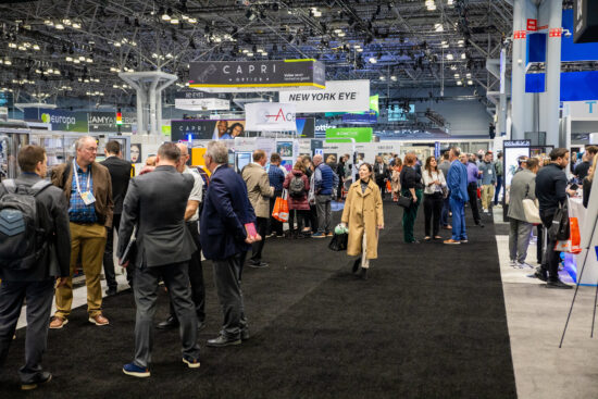 A busy eyewear trade show floor with crowds of people walking between branded exhibit booths like Capri Optics and New York Eye.