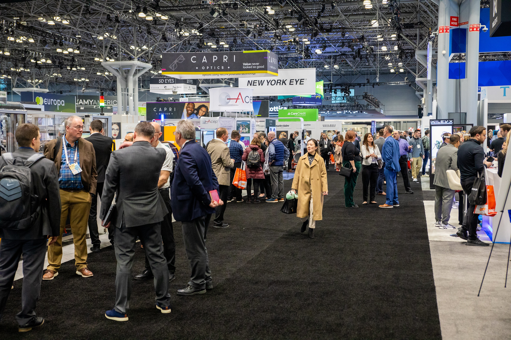 A busy eyewear trade show floor with crowds of people walking between branded exhibit booths like Capri Optics and New York Eye.