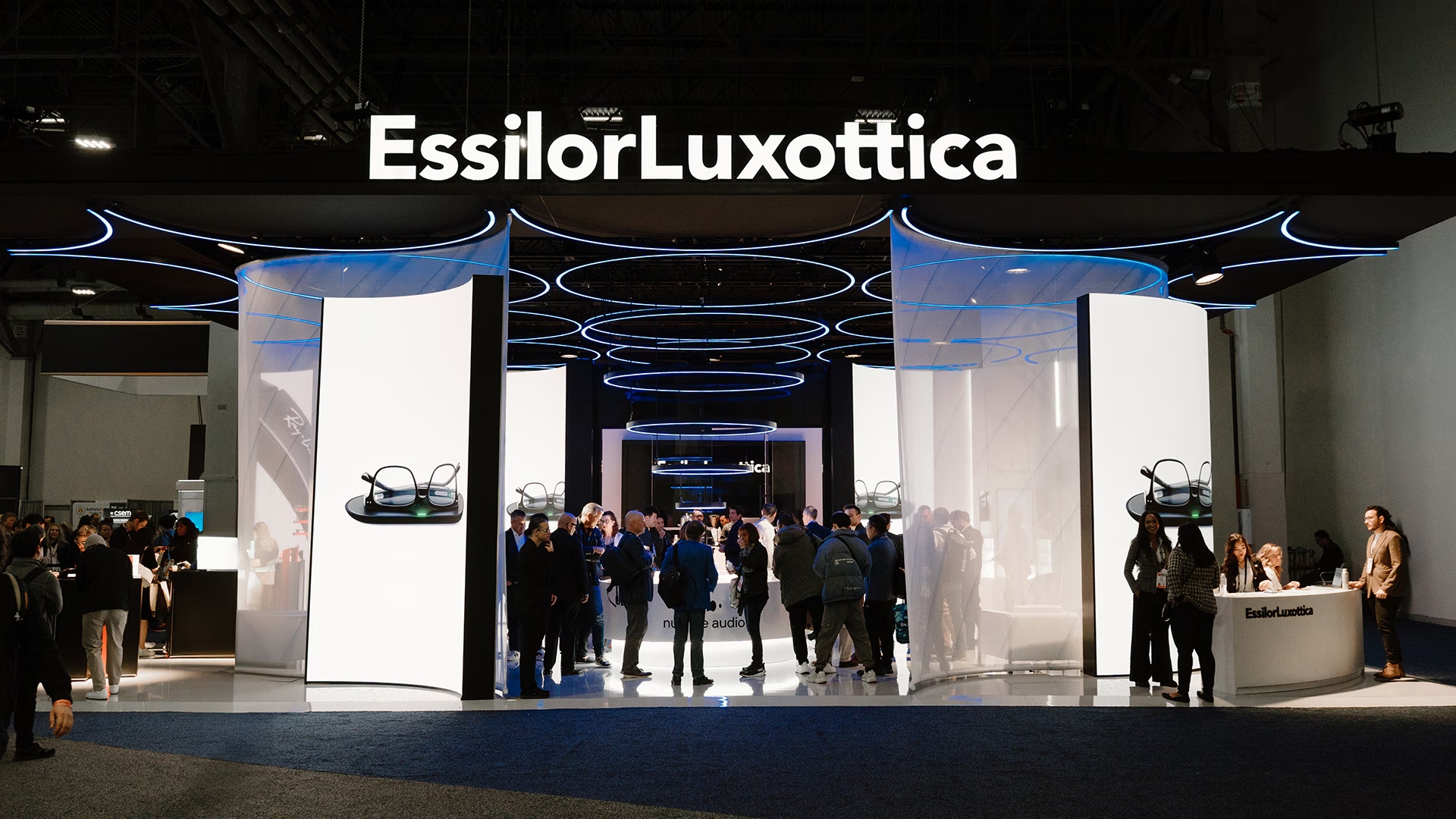 A wide shot of the modern EssilorLuxottica exhibition booth featuring large digital displays of smart eyewear and a crowd of visitors.