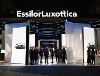 Vue d'ensemble du stand d'exposition moderne d'EssilorLuxottica avec de grands écrans numériques présentant des lunettes intelligentes et de nombreux visiteurs.