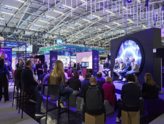 An audience at opti 2026 watches a panel discussion on a modern stage with a large circular screen, surrounded by trade show booths.