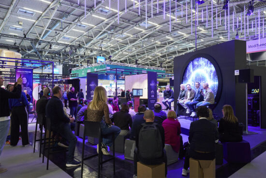 An audience at opti 2026 watches a panel discussion on a modern stage with a large circular screen, surrounded by trade show booths.