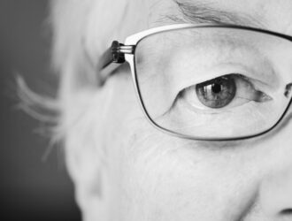 Gros plan en noir et blanc de l'œil d'une personne âgée derrière des lunettes à monture fine, mettant l'accent sur la santé visuelle.